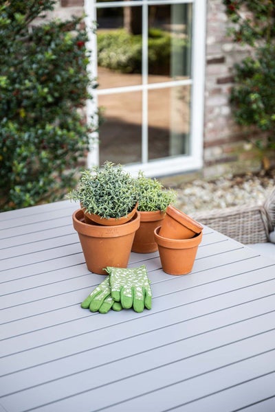 Terracotta-Blumentöpfe mit Pflanzen und ein Paar grüne Gartenhandschuhe auf einem hellgrauen Gartentisch im Außenbereich.
