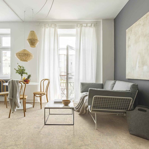 Hell gestaltetes Wohnzimmer mit strukturiertem Bodenbelag, grauem Sofa, Esstisch und Bambus-Pendelleuchten vor einem Fenster mit weißen Vorhängen.