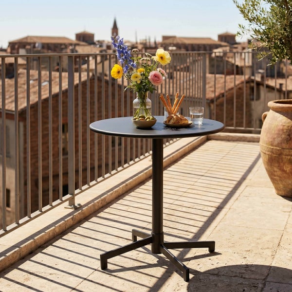 Runder Metall-Bistrotisch auf einem Balkon mit Steinboden und Stadtblick, dekoriert mit Blumen und Snacks.