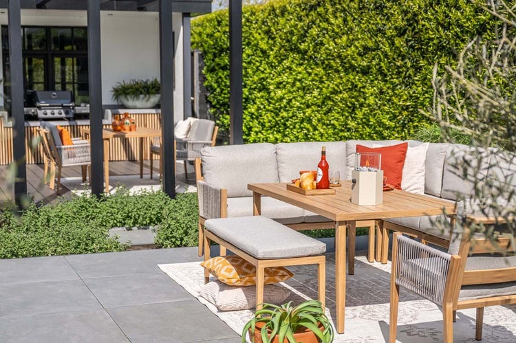 Garten Lounge Essgruppe aus Holz mit Ecksofa, grauen Polstern, Holztisch, Sitzbank und Sessel auf einer hellen Terrasse mit grüner Hecke.