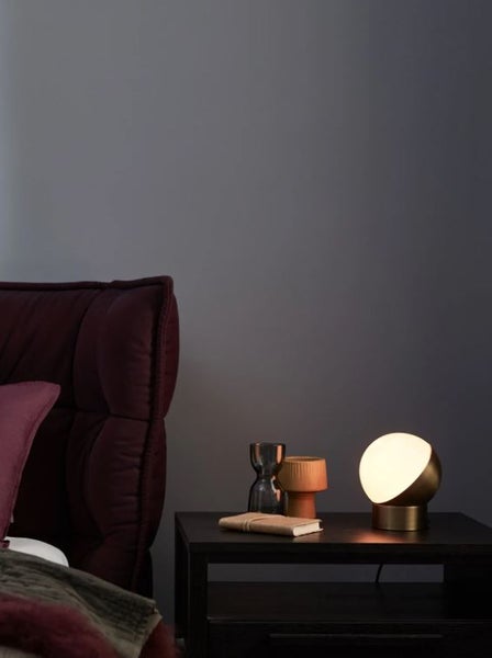 Moderne runde Tischleuchte in Messingoptik auf einem Nachttisch neben einem Bett mit Dekoration.