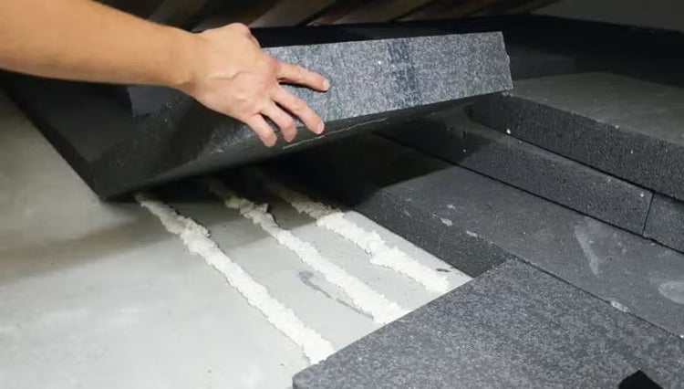Eine Person verlegt dunkelgraue Dämmplatten auf einem Betonboden mit Hilfe von weißem Klebeschaum.