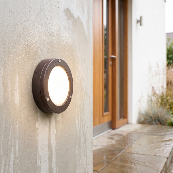 Helle, runde Außenwandleuchte aus Metall und Kunststoff an einer verputzten Wand im Außenbereich neben einer Holztür und Steinplatten.