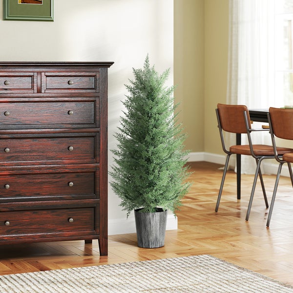 Künstliche Pflanze in Form einer Zypresse im Topf neben einer dunklen Holzkommode in einem hellen, organisierten Zimmer mit Parkettboden.