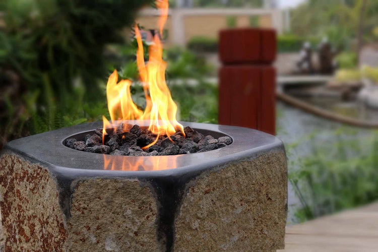 Gas-Feuerstelle mit einer Außenseite aus Naturstein, glatter Oberseite und orangefarbenen Flammen aus schwarzen Lavasteinen.