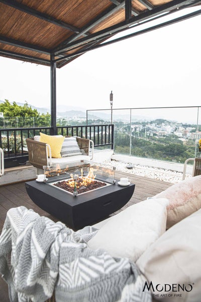 Moderne Gas-Feuerstelle auf einer Dachterrasse mit Loungemöbeln, Holzdeck, Glasgeländer und Blick auf die Stadt. Modeno Logo.