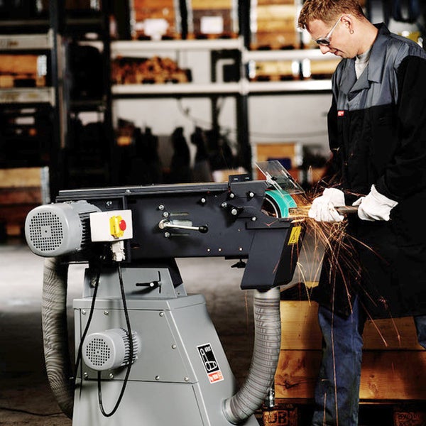 Handwerker schleift Metall an einer stationären Bandschleifmaschine in einer hellen Werkstatt, Funkenflug und Schutzkleidung.