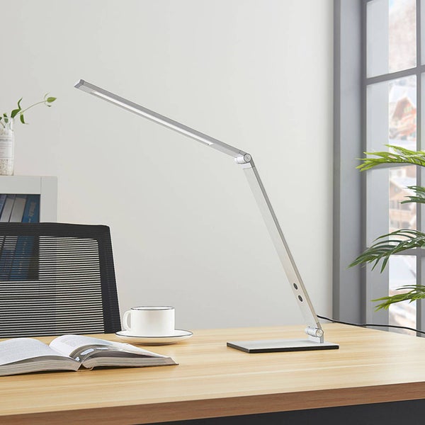 Moderne silberne LED-Schreibtischlampe mit verstellbarem Arm auf einem Holzschreibtisch mit Buch und Kaffeetasse in einem hellen Büro.