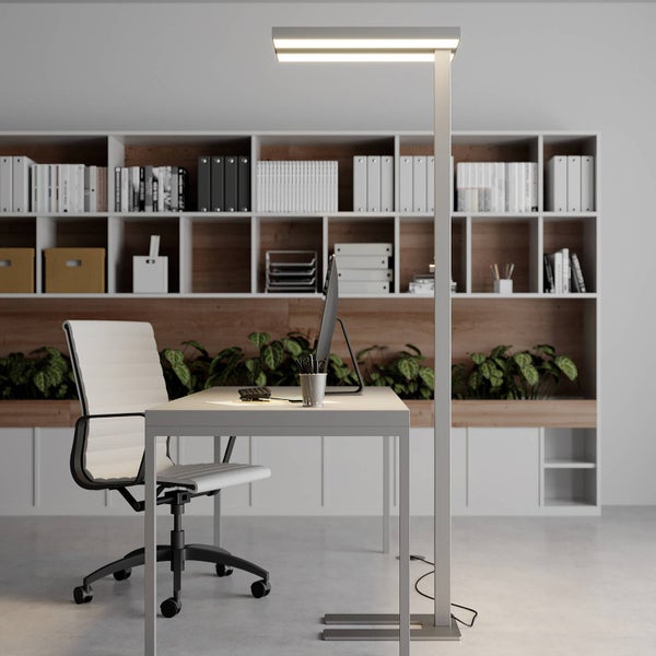 Modernes, helles Büro mit weißem Schreibtisch, ergonomischem Bürostuhl und einer schlanken Stehleuchte vor einem Regal mit Büchern und Pflanzen.