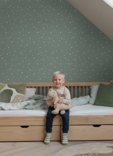 Kinderzimmer mit Sternentapete an einer Wand mit Dachschräge. Ein Kind sitzt auf einem Holzbett mit Schubladen und hält ein Stofftier.