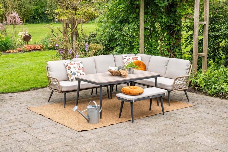 Garten-Lounge-Set aus Metall mit Ecksofa, Esstisch und Bank mit beigen Auflagen auf einer Terrasse mit Teppich in einem Garten.
