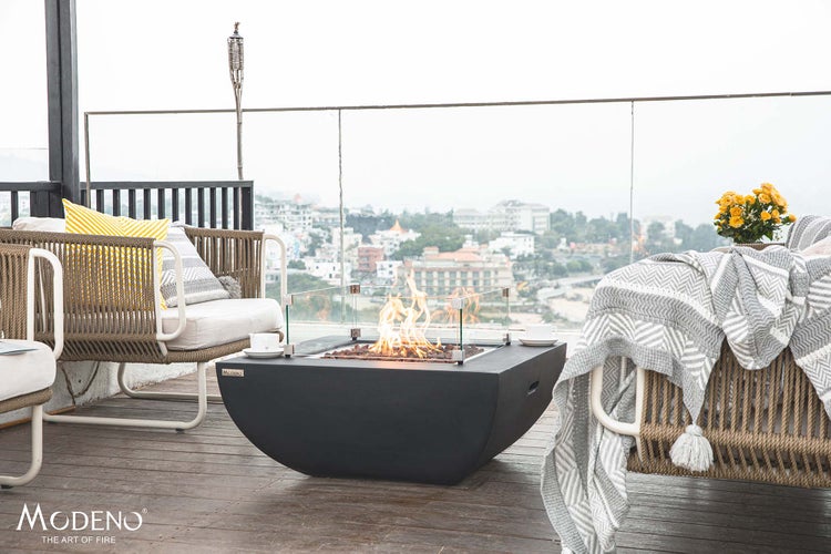 Modeno Gas-Feuerstelle auf einer modernen Terrasse mit Loungesesseln, Glaswindschutz und Blick auf die Stadt.