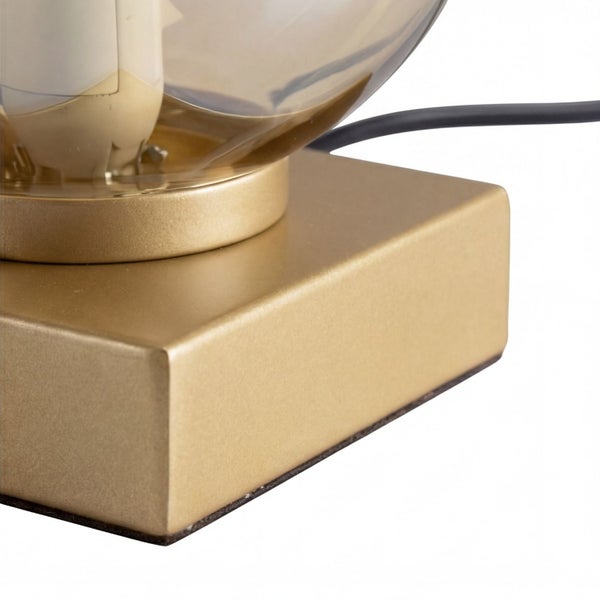 Detail einer quadratischen goldfarbenen Lampenbasis aus Metall mit kreisförmiger Halterung und getöntem Glasschirm.