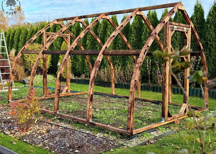 Holzskelett eines bogenförmigen Gewächshauses im Garten mit dunklen geschwungenen Rippen und Türrahmen. Koks Logo.