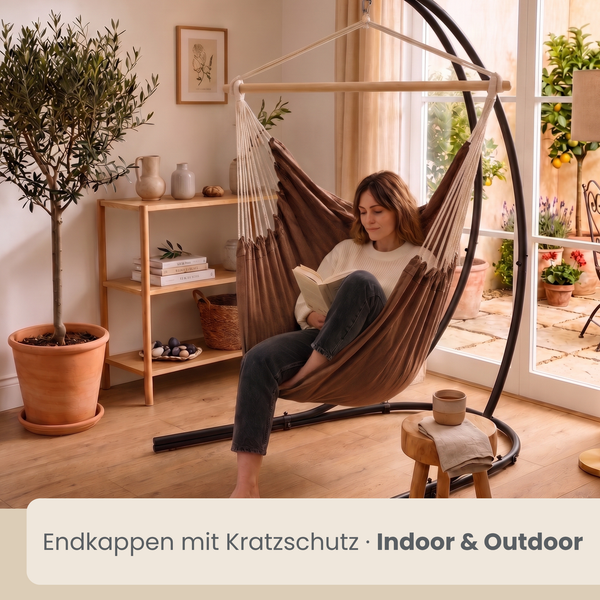 Eine Person liest entspannt in einem Hängesessel mit Gestell in einem hellen, wohnlich eingerichteten Raum mit Terrassenzugang und Pflanzen.