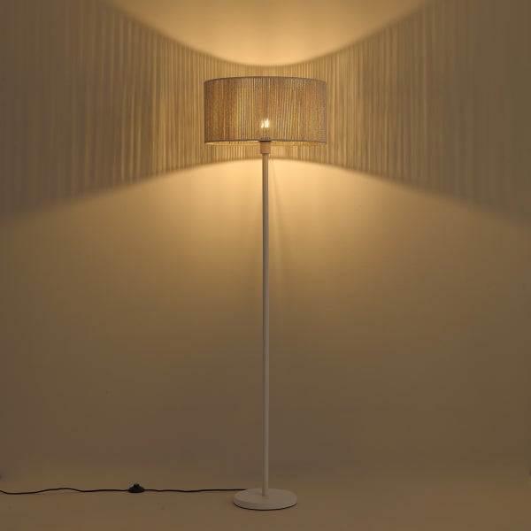 Eingeschaltete Stehlampe mit rundem Fadenschirm, die ein warmes Licht und gestreifte Schatten an die Wand wirft.