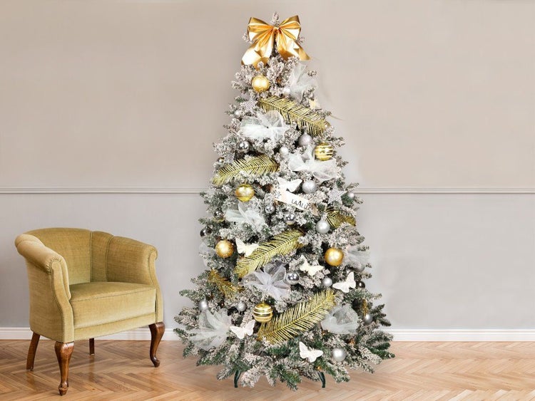 Geschmückter künstlicher Weihnachtsbaum mit Schneeoptik, dekoriert mit Kugeln, Schleifen und goldenen Zweigen neben einem Sessel.