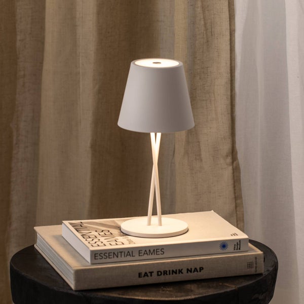 Moderne Tischleuchte mit konischem Schirm und schmalen Beinen, platziert auf Büchern vor einem Vorhang.