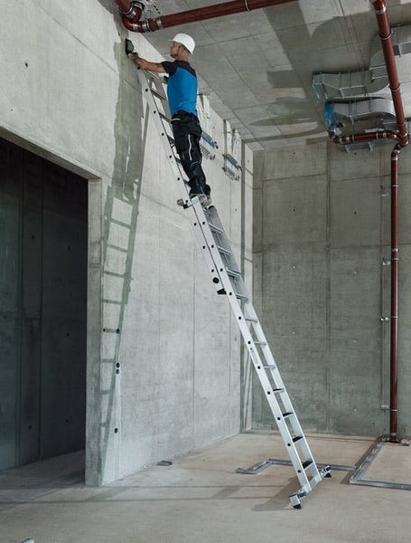 Handwerker mit Schutzhelm steht auf einer langen Aluminium Mehrzweckleiter an einer Betonwand in einem Rohbau.