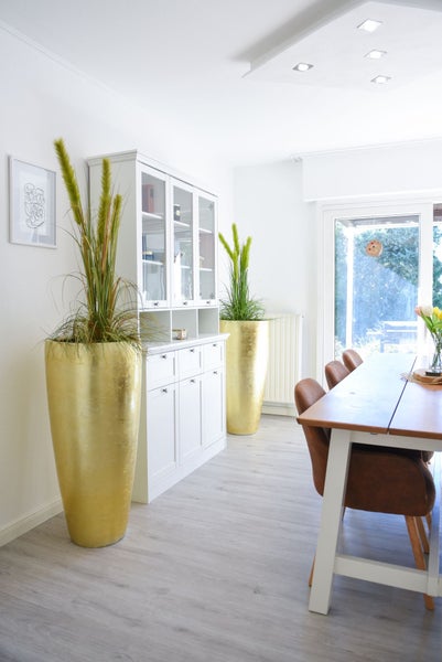 Helles Esszimmer mit weißem Buffetschrank, zwei hohen goldenen Pflanzgefäßen mit Kunstgras und einem Esstisch aus Holz mit braunen Stühlen.