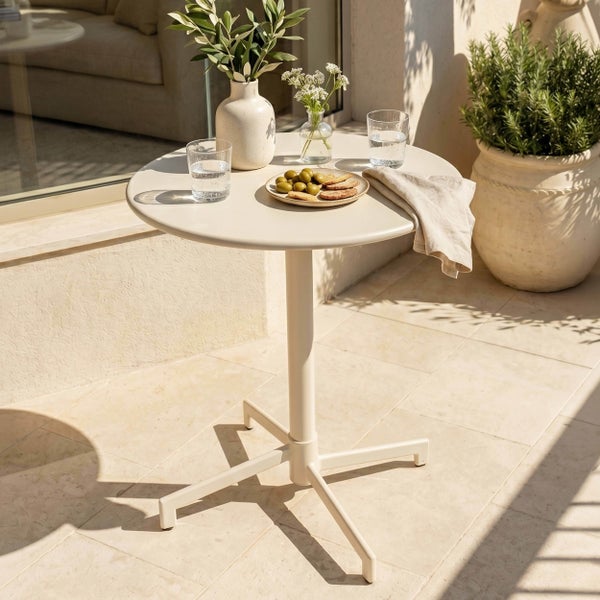 Runder Gartentisch aus Metall auf einer hellen Terrasse, dekoriert mit Vasen, Gläsern und einem Teller mit Oliven.