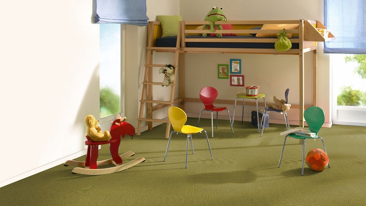 Helles Kinderzimmer mit einem Hochbett aus Holz, bunten Stühlen und einem Schaukelelk auf einem Boden aus grünen Teppichfliesen.