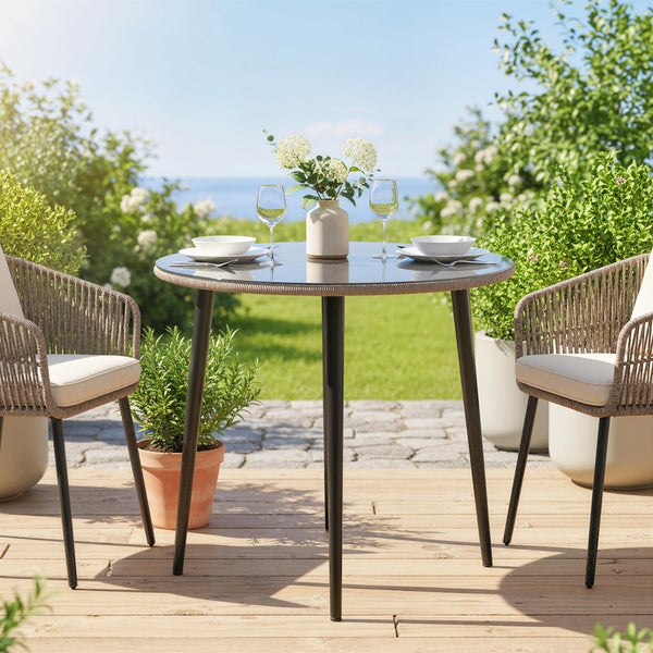 Moderner runder Gartentisch mit Glasplatte und zwei Seilgeflecht-Stühlen mit Polstern auf einer Holzterrasse vor Garten- und Meereskulisse.
