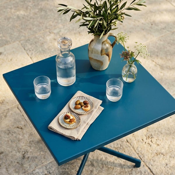 Quadratischer Bistro-Tisch aus Metall auf einer Steinterrasse, gedeckt mit Gläsern, einer Karaffe, Snacktellern und Vasen mit Olivenzweigen.