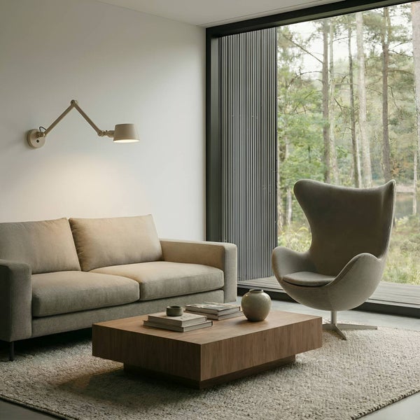 Wohnzimmer mit Sofa, Sessel, Couchtisch, Teppich und Wandlampe vor einem Fenster mit Blick auf Bäume