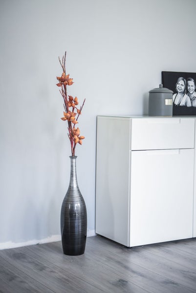 Schlanke Bodenvase mit Querstreifen und Trockenzweigen mit orangefarbenen Kapseln neben einer weißen Kommode in einem hellen Zimmer.