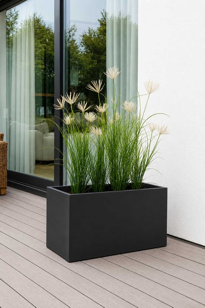 Rechteckiger, dunkler Pflanzkübel mit künstlichem Ziergras auf einer modernen Terrasse mit Holzdielen vor einer Glasfront.