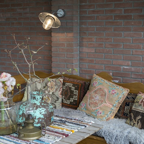Eingeschaltete Außenwandleuchte mit Glasschirm an einer Klinkerwand über einer Sitzecke mit Holzbank, bunten Kissen und Dekoration.