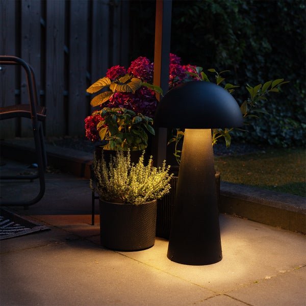 Moderne pilzförmige Außenleuchte auf einer Steinterrasse neben Blumentöpfen mit violetten Blüten bei Dämmerung.