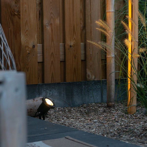 Schwarzer Gartenstrahler mit Erdspieß beleuchtet Holzzaun in einem Kiesbeet neben Ziergras und Bambusstäben.