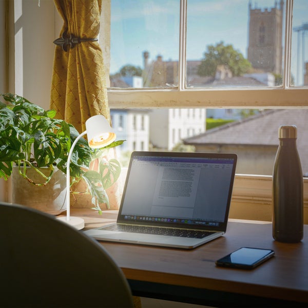 Weiße LED Schreibtischleuchte mit flexiblem Hals auf einem Holztisch neben einem Laptop und einer Pflanze vor einem hellen Fenster.