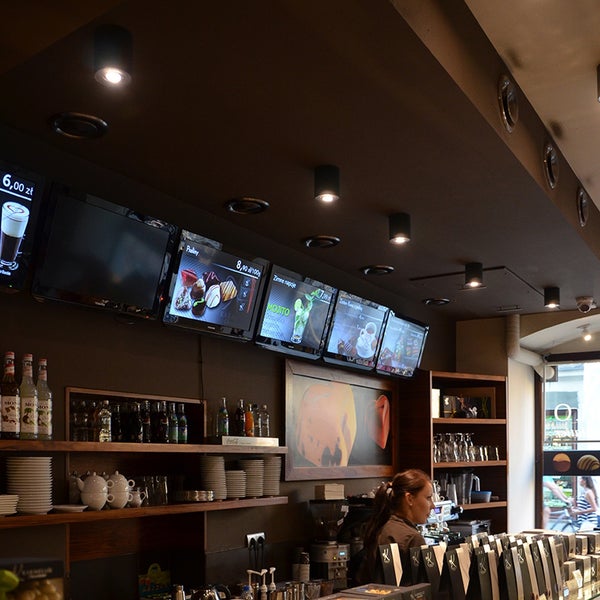 Innenaufnahme eines Cafés mit Deckenleuchten, Monitoren mit Menüanzeigen und Regalen mit Getränken und Geschirr