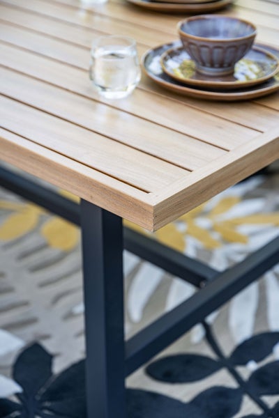 Gartentisch mit Holzplatte und Metallgestell. Detailaufnahme der Tischecke mit Geschirr und Glas.