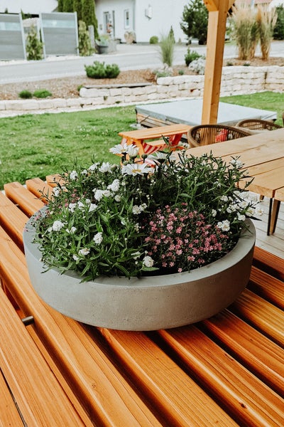 Rundes Pflanzgefäß in Betonoptik mit weißen und rosa Blumen auf einem hölzernen Gartentisch im Außenbereich.