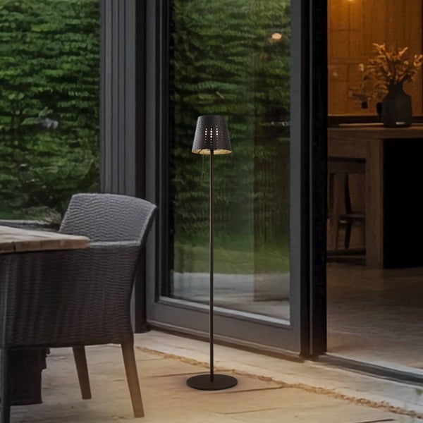 Moderne Außenstehleuchte aus Metall mit gelochtem Schirm auf einer Terrasse neben einem Korbstuhl und einer Glastür.