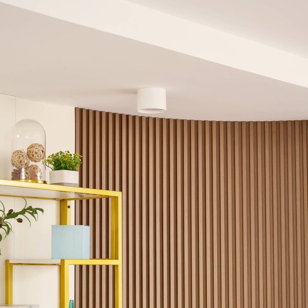 Kleiner weißer zylindrischer Deckenaufbaustrahler in einem Raum mit vertikaler Holzlamellenwand und einem goldenen Regal.