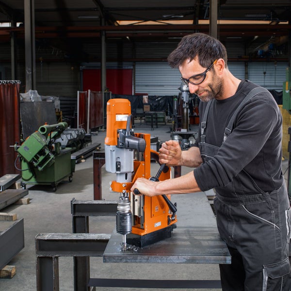 Ein Handwerker in Arbeitskleidung und Schutzbrille bedient eine Magnetkernbohrmaschine auf einer Metallplatte in einer Industriehalle.