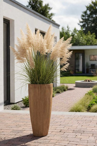 Hoher, konischer Pflanzkübel in Holzoptik mit Pampasgras auf einer modernen Terrasse mit Pflastersteinen und Garten im Hintergrund.