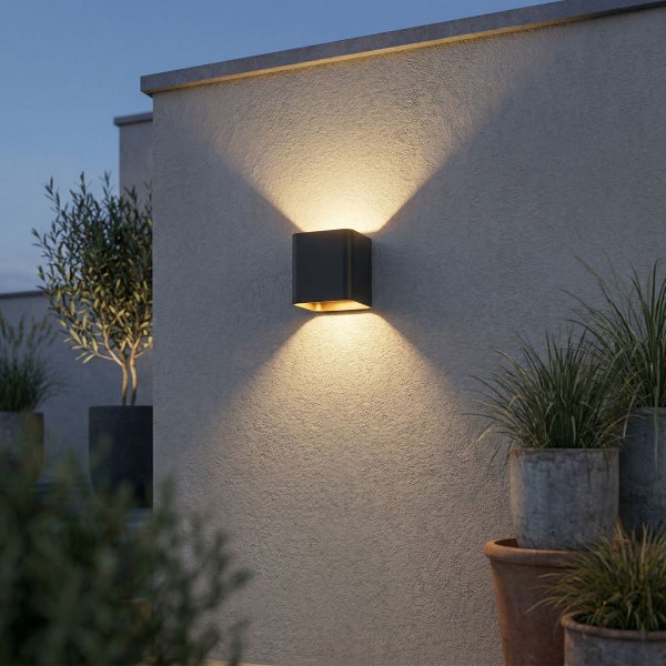 Quadratische Außenwandleuchte mit Lichtkegeln nach oben und unten an einer strukturierten Außenwand mit Kübelpflanzen.