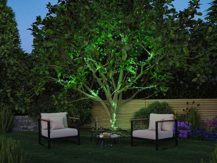 Grüne Gartenbeleuchtung an einem Baum in einem Sitzbereich mit zwei Loungesesseln, Tischen und einem Holzzaun bei Nacht.