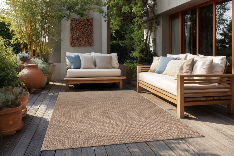 Ein gewebter Outdoor-Teppich mit Fischgrätmuster auf einer Holzterrasse zwischen zwei Loungesofas, umgeben von Pflanzen und Töpfen.