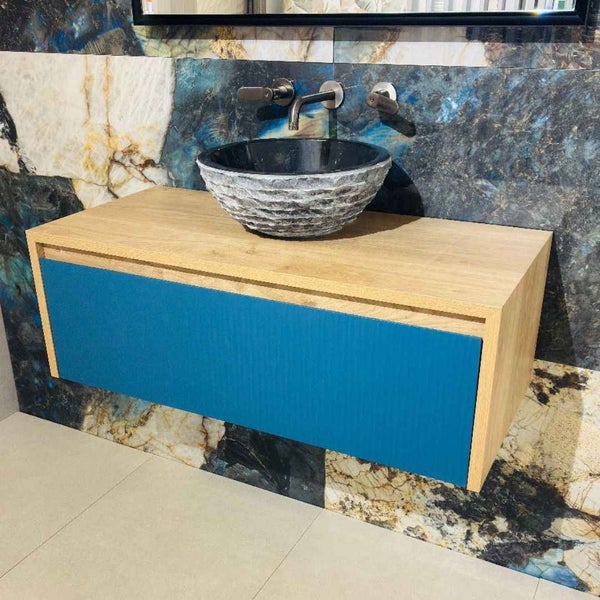 Moderner Waschtischunterschrank aus Holz mit blauer Front, rundem Naturstein-Aufsatzwaschbecken und Wandarmatur vor marmorierten Wandfliesen.