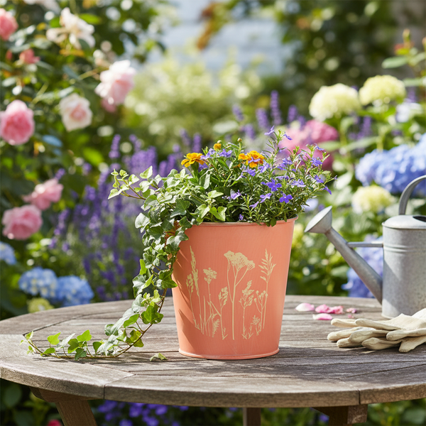 Runder Pflanztopf aus Metall mit Naturmotiv auf Holztisch mit Efeu, Gartenhandschuhen und Gießkanne in einem hellen, organisierten Garten.