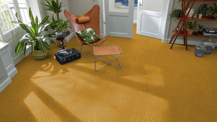 Heller Wohnraum mit strukturierten gelben Teppichfliesen, einem ledernen Butterfly-Sessel, Zimmerpflanzen und einem Holzregal.