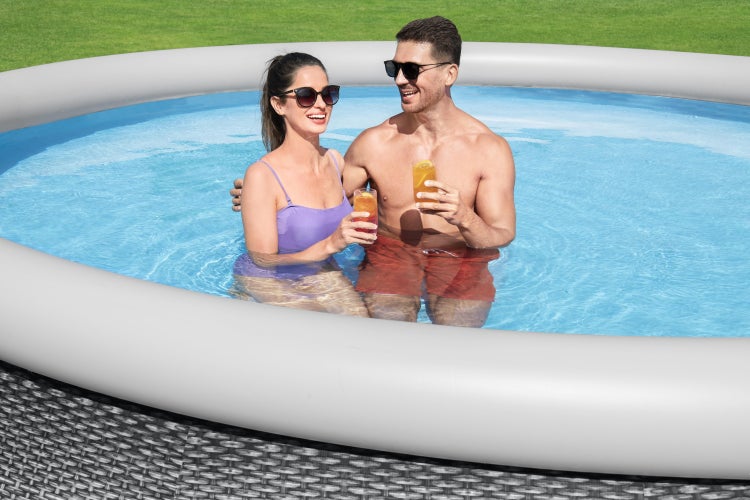 Ein Mann und eine Frau entspannen in einem aufblasbaren Gartenpool mit Rattan-Dekor und grauem Luftring auf einer Rasenfläche.