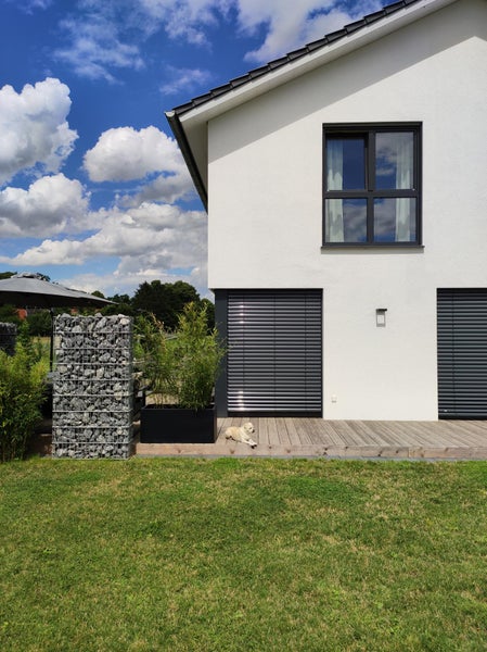 Moderne Hausfassade mit Außenjalousien, einer Holzterrasse, einer Gabione und einer Rasenfläche.